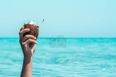 Foto de Primer plano de la mano de una persona irreconocible sosteniendo la mitad de la cáscara de coco con rodajas de coco en ella contra el mar y el cielo. Mano mojada en verano con cáscara de coco. Mano levantada sosteniendo coco contra el mar - Imagen libre de derechos