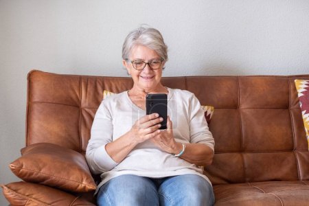 Foto de Mujer mayor sentada en el sofá y leyendo mensajes de texto en el teléfono móvil en casa. Mujer mayor en gafas de surf y el uso de las redes sociales en el teléfono móvil en la sala de estar. - Imagen libre de derechos