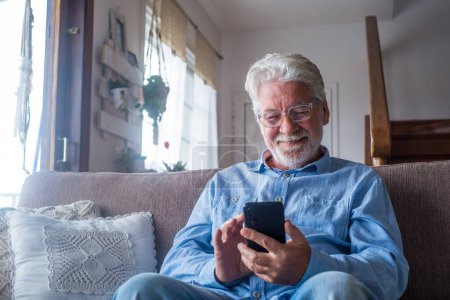 Foto de Viejo sonriendo sentado en el sofá en la sala de estar sosteniendo el teléfono, disfrutando de usar el teléfono inteligente sintiéndose satisfecho enviando mensajes, llamando a amigos, navegando por el concepto en línea web - Imagen libre de derechos