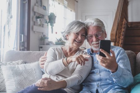 Lächelnd aufrichtiges älteres verheiratetes Familienpaar, das Handy-Videotelefonie mit Freunden führt, die distanzierte Kommunikation mit erwachsenen Kindern genießt, Smartphone-Anwendungen zu Hause nutzt.  