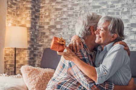 Portrait of two cute and old seniors at home having fun together. Mature man  giving a gift at his wife for Christmas or anniversary. Surprised pensioner woman hugging his husband. 