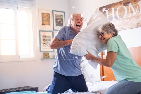 Foto de Pareja de dos personas mayores felices divirtiéndose jugando juntos en la cama en casa luchando con almohadas disfrutando - almohadas guerra en el interior por la mañana - Imagen libre de derechos
