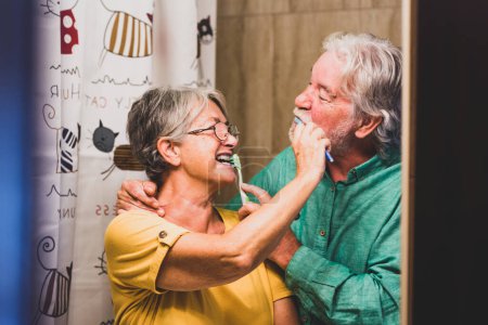 pareja de dos ancianos felices y sonrientes cepillándose los dientes el uno al otro juntos en casa en el baño - autocuidado y cuidar de sí mismos 