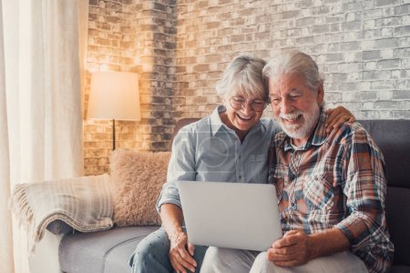 Linda pareja de ancianos sentados en el sofá utilizando el ordenador portátil juntos de compras y navegar por la red. Dos personas maduras en el salón disfrutando de la tecnología. Retrato de seniors riendo en el amor. 