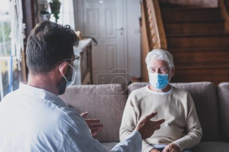 Photo for Male professional doctor consulting senior patient during medical care visit wearing masks. Young man physician and old mature senior talking providing medical assistance sitting on sofa. Elderly people home care. Covid and coronavirus positive test - Royalty Free Image