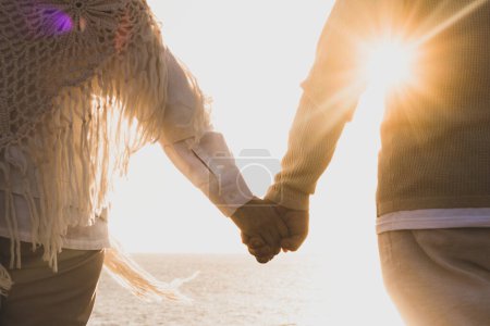 Foto de Pareja de dos personas mayores felices y activas divirtiéndose y disfrutando juntos del verano en la playa caminando tomados de la mano con la puesta de sol al fondo - Imagen libre de derechos