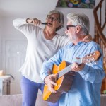 Couple of two happy seniors or mature and old people singing and dancing together at home indoor. Retired man playing the guitar while his wife is singing with a remote control of TV. 