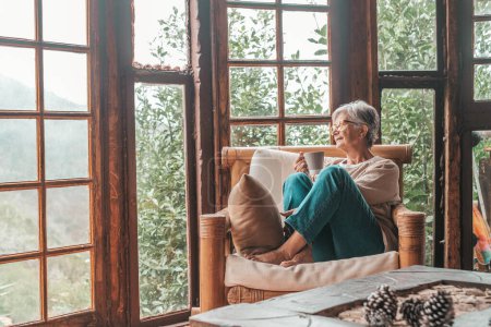 Photo for Senior woman sitting on sofa and holding coffee mug while looking away at home. Elderly woman relaxing on couch in living room. Thoughtful woman having coffee on sofa at home - Royalty Free Image
