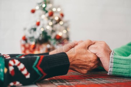 Couple aîné en vêtements chauds se tenant les mains devant l'arbre de Noël décoré à la maison. Aimer vieux couple hétérosexuel romantique célébrant Noël festival ensemble 
