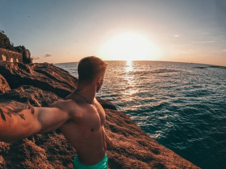 Ein fröhlicher junger Mann, der allein am Strand spaziert, mit Sonnenuntergang im Hintergrund. Männliche Person hat Spaß und genießt die Freiheit, Selfie zu machen.