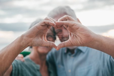 Foto de Close up portrait happy sincere middle aged elderly retired family couple making heart gesture with fingers, showing love or demonstrating sincere feelings together outdoors, looking at camera. - Imagen libre de derechos