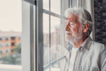 Téléchargez les photos : Heureux homme réfléchi âgé de 70 ans regardant par la fenêtre loin avec espoir, pensant à une bonne santé, retraite, prestations d'assurance, rêver d'avenir. Pensionné âgé en attente d'une réunion avec sa famille - en image libre de droit