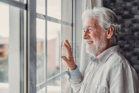 Téléchargez les photos : Heureux homme réfléchi âgé de 70 ans regardant par la fenêtre loin avec espoir, pensant à une bonne santé, retraite, prestations d'assurance, rêver d'avenir. Pensionné âgé en attente d'une réunion avec sa famille - en image libre de droit