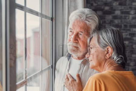 Téléchargez les photos : Pensive elderly mature senior man in eyeglasses looking in distance out of window, thinking of personal problems. Old woman wife consoling and hugging sad husband, copy space - en image libre de droit