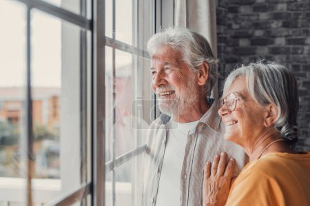 Happy bonding loving middle aged senior retired couple standing near window, looking in distance, recollecting good memories or planning common future, enjoying peaceful moment together at home.