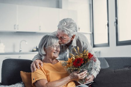 Téléchargez les photos : Vieil homme donnant des fleurs à sa femme assise sur le canapé à la maison pour la Saint-Valentin. Les retraités apprécient la surprise ensemble. Dans l'amour les gens s'amusent - en image libre de droit