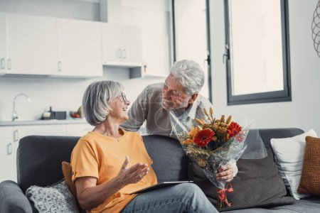 Photo for Old man giving flowers at his wife sitting on the sofa at home for the San Valentines day. Pensioners enjoying surprise together. In love people having fun - Royalty Free Image