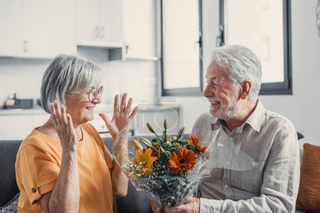 Photo for Old man giving flowers at his wife sitting on the sofa at home for the San Valentines day. Pensioners enjoying surprise together. In love people having fun - Royalty Free Image