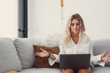 Téléchargez les photos : Portrait d'une jeune femme blonde bouleversée et malheureuse utilisant un ordinateur portable sur le canapé ayant des difficultés à travailler et à étudier au travail à la maison. - en image libre de droit