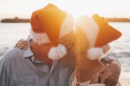 Foto de Viejos linda pareja de personas maduras disfrutando y divirtiéndose juntos en la playa con sombreros de Navidad en los días de vacaciones. Abrazado en la playa con el atardecer al fondo en invierno. - Imagen libre de derechos