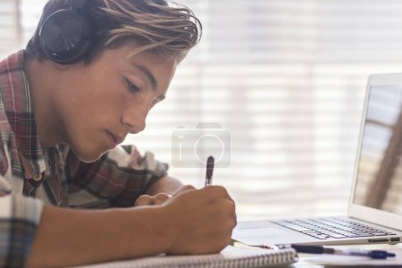 kaukasischer Teenager drinnen Hausaufgaben auf dem Tisch zu Hause machen - blonder Kerl schreibt und liest in seinem Laptop oder Computer, um die besten Noten zu bekommen