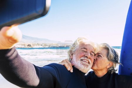 schönes Paar von zwei Senioren am Strand mit Neoprenanzug und Surfbrett, die ein Selfie machen, bevor sie zusammen surfen gehen - aktive reife Menschen