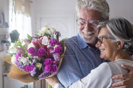 Téléchargez les photos : Portrait de couple de deux personnes âgées heureuses et amoureuses ou de personnes âgées et matures tenant des fleurs à la maison regardant dehors. Retraités adultes profitant et célébrant ensemble des vacances. - en image libre de droit
