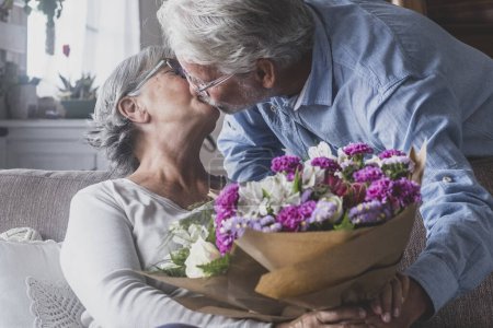 Téléchargez les photos : Vieil homme donnant des fleurs à sa femme assise sur le canapé à la maison pour la Saint-Valentin. Les retraités aiment s'embrasser par surprise ensemble. Dans l'amour les gens s'amusent. - en image libre de droit