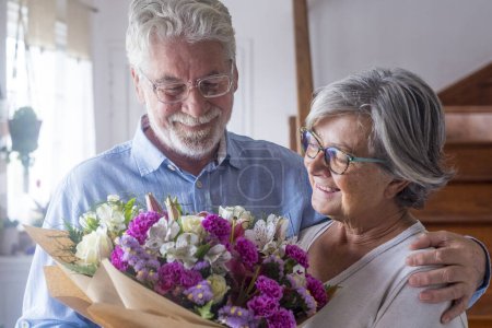 Photo for Portrait of couple of two happy and in love seniors or mature and old people holding flowers at home looking at the camera. Pensioners adult enjoying and celebrating holiday together. - Royalty Free Image