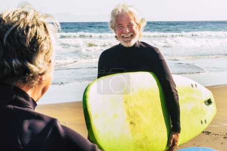 Ein paar Senioren am Strand mit schwarzen Neoprenanzügen, die ein Surfgerät in der Hand halten, um am Strand zu surfen - aktive reife und pensionierte Menschen, die in ihrem Urlaub oder ihrer Freizeit gemeinsam fröhliche Aktivitäten ausüben