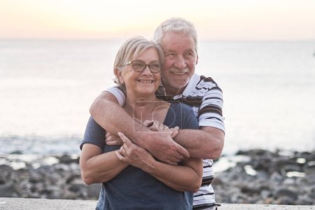 couple of seniors hogging at the beach with a lot of love - retired together - woman with glasses and man with sea background