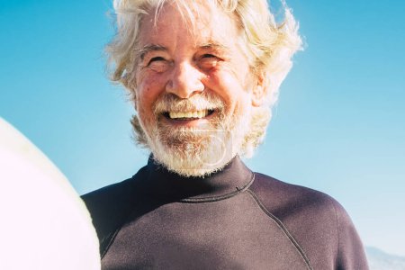 glücklicher älterer Mann mit schwarzem Neoprenanzug und surfbar am Strand bereit zum Surfen - Nahaufnahme von Senior lächelnd und lachend mit dem Meer oder Ozean im Hintergrund