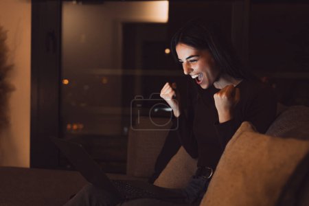 Foto de Mujer sentarse en el sofá leer e-mail en el ordenador portátil hace que sí gesto se siente feliz. Empresaria obtener grandes noticias de negocios, celebrar el crecimiento de la carrera, avanzar. Logro, victoria, momento de subasta victoria en la noche - Imagen libre de derechos