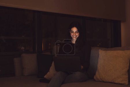 Foto de Primer plano de una joven feliz alegre mujer sonriendo y el uso de ordenador portátil en casa sentado en el sofá de trabajo y estudiar solo en la noche. Millennial chica surfeando la red interior con pantalla de luz en su cara relajante en el sofá. - Imagen libre de derechos