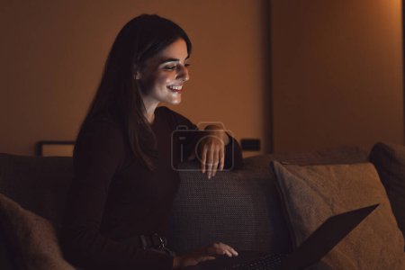 Photo for Close up of one young happy cheerful woman smiling and using laptop computer at home sitting on sofa working and studying alone at late night. Millennial girl surfing the net indoor with light screen on her face relaxing on couch. - Royalty Free Image