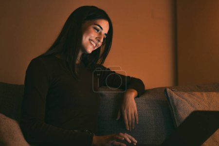 Téléchargez les photos : Gros plan d'une jeune femme joyeuse et souriante qui utilise un ordinateur portable à la maison, assise sur un canapé, travaillant et étudiant seule tard dans la nuit. Millennial fille surfer sur le filet intérieur avec écran lumineux sur son visage relaxant sur le canapé. - en image libre de droit