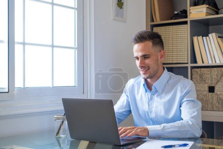 Téléchargez les photos : Heureux jeune homme d'affaires millénaire caucasien travaillant à la maison au bureau avec ordinateur portable ou ordinateur s'amuser. Jeune homme en vidéoconférence prenant soin de l'entreprise ou du marché. - en image libre de droit
