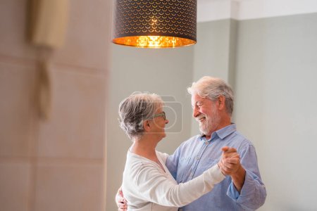 Téléchargez les photos : Joyeux vieux couple romantique retraité actif dansant en riant dans le salon, heureuse épouse d'âge moyen et mari aîné s'amusant à la maison, souriant grands-parents de la famille aînée se relaxant liens ensemble - en image libre de droit