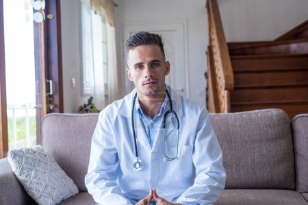 Photo for Portrait of smiling caucasian young male doctor at home in white medical uniform and stethoscope sit on the sofa look at camera, happy positive man GP or physician posing at workplace - Royalty Free Image
