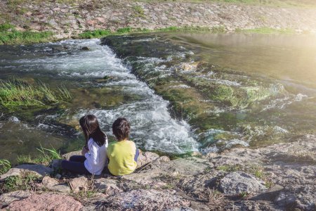 Téléchargez les photos : Des enfants assis au bord de la rivière. Deux enfants ou frères et sœurs regardant l'écoulement rapide de l'eau et jouant à l'extérieur. remblai de pierre, explorer le concept du monde. - en image libre de droit
