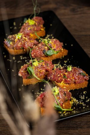 Photo for Beef tartare garnished with microgreens and a slice of avocado, topped with toasted buckwheat bread sauteed in garlic butter. Food lies on a rectangular ceramic plate on a wooden background. - Royalty Free Image