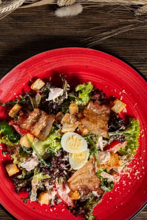 Photo for Caesar salad with breaded chicken and grilled bacon in a red ceramic plate on a dark wooden background. - Royalty Free Image
