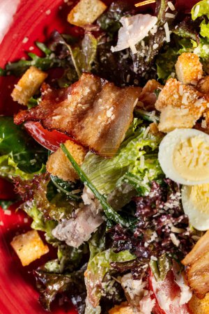 Téléchargez les photos : Salade César au poulet pané et bacon grillé dans une assiette en céramique rouge sur fond de bois foncé. - en image libre de droit