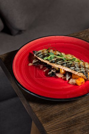 Photo for Grilled mackerel fillet with mustard sauce and garnished with microgreens lies on oven-baked vegetables. Food lies on a red ceramic plate on a dark, wooden background. - Royalty Free Image