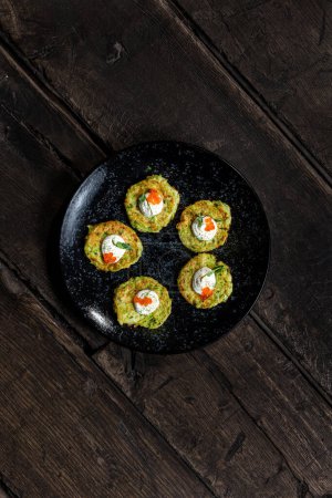 Photo for Zucchini fritters with sour cream and red caviar garnished with green onion pieces on a dark ceramic plate. The plate stands on dark wooden boards. - Royalty Free Image