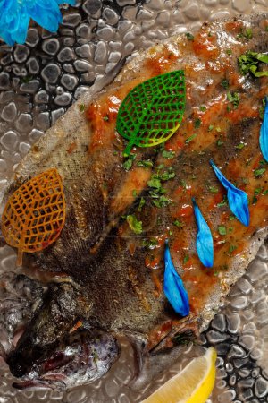 Téléchargez les photos : Truite de rivière grillée à la tomate et sauce aux légumes à l'ail. Le poisson repose sur une plaque de verre transparente sur un fond clair de blocs de bois. - en image libre de droit