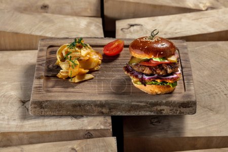 Photo for Cheeseburger with beef patty and chicken patty, lettuce, goat cheese and fresh tomatoes with mustard sauce. Nearby lies fried potatoes and pickled tomato. Food lies on a wooden stand on a light background. - Royalty Free Image