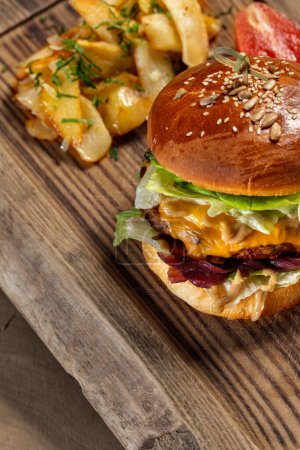 Photo for Cheeseburger with duck patty, lettuce, red onion, cheese and fresh tomatoes with mustard sauce. Nearby lies fried potatoes and pickled tomato. Food lies on a wooden stand on a light background. - Royalty Free Image