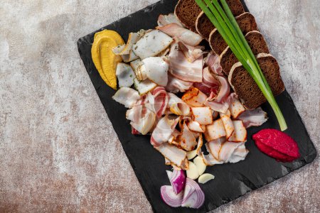 Photo for A set of meat snacks from bacon, smoked and baked meat. green onions, black bread toast with mustard and horseradish in beetroot on a black slate plate. The plate stands on a light background. - Royalty Free Image