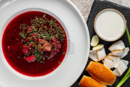 Ukrainian borscht soup with garlic donuts, green onions and bacon, next to it is a bowl of sour cream. The soup is poured into a light ceramic plate. Dishes stand on a light background.